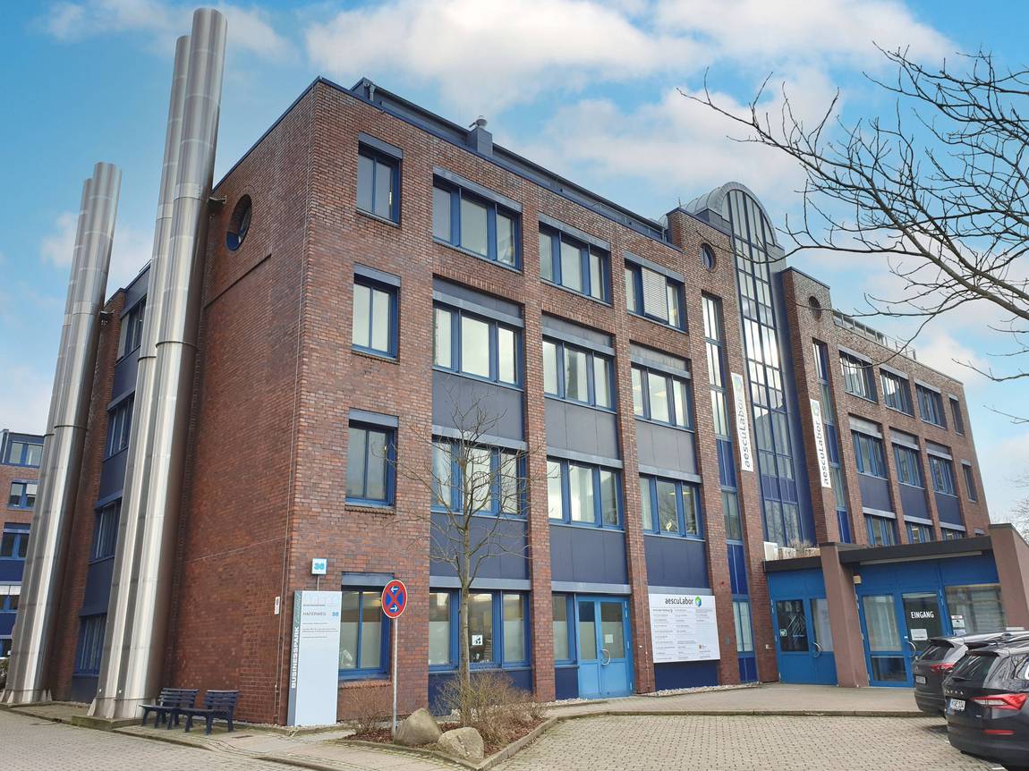 Ein modernes Bürogebäude aus rotem Backstein mit großen Fenstern, einem zentralen Glasbogen und hohen silbernen Schornsteinen, unter einem blauen Himmel mit vereinzelten Wolken. Autos sind geparkt und ein blattloser Baum ist davor zu sehen.
