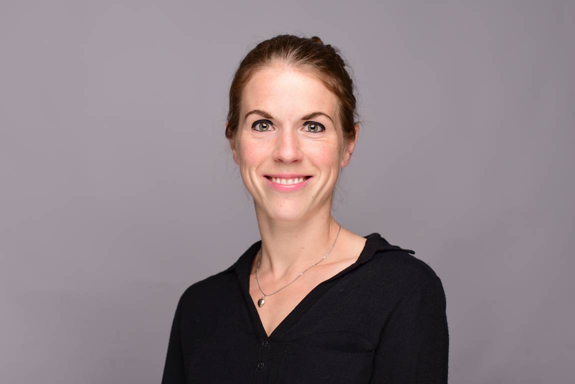 Eine Frau mit heller Haut und zurückgebundenen braunen Haaren lächelt in die Kamera. Sie trägt eine schwarze Bluse und eine Halskette und steht vor einem schlichten grauen Hintergrund.